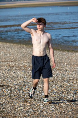 Teenage boy walking towards the camera and shielding his eyes from the sun, along a beach on warm summer's day
