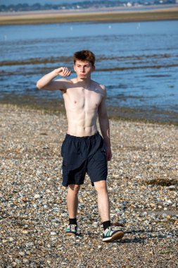Teenage boy walking towards the camera and shielding his eyes from the sun, along a beach on warm summer's day