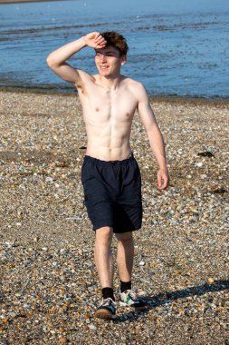 Teenage boy walking towards the camera and shielding his eyes from the sun, along a beach on warm summer's day