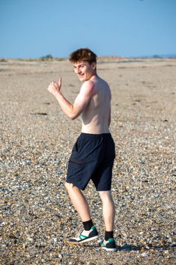 Teenage boy turning  towards the camera, along a beach on warm summer's day, giving a thumbs u