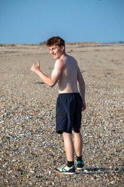 Teenage boy turning  towards the camera, along a beach on warm summer's day, giving a thumbs u
