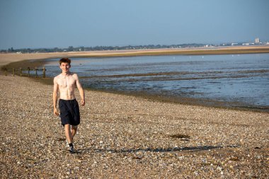 Teenage boy walking towards the camera, along a beach on warm summer's day, landscape shot
