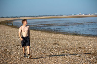 Teenage boy walking towards the camera, along a beach on warm summer's day, landscape shot