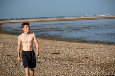 Teenage boy walking towards the camera, along a beach on warm summer's day, landscape shot