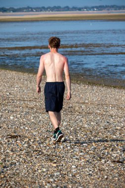 Teenage boy walking away from the camera, along a beach on warm summer's da
