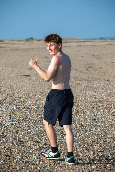 Teenage boy turning  towards the camera, along a beach on warm summer's day, giving a thumbs u