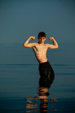 Tişörtsüz, kaslı ergen çocuk ılık bir yaz gününde gün batımında denizde kaslarını esnetiyor.