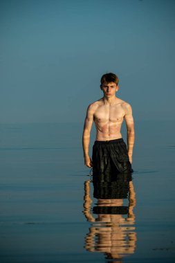 Tişörtsüz, kaslı ergen çocuk ılık bir yaz gününde gün batımında denizde kaslarını esnetiyor.