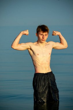 Tişörtsüz, kaslı ergen çocuk ılık bir yaz gününde gün batımında denizde kaslarını esnetiyor.