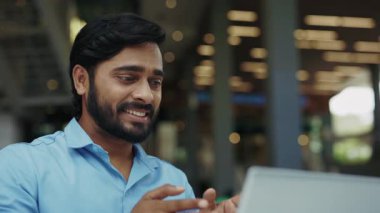 Caddedeki kafede dizüstü bilgisayarla çekilmiş mutlu Hintli girişimci jestlerinin portresi. Neşeli adam online toplantı sırasında şirket ofisinin dışında cihaz kullanarak işaret dili kullanıyor.