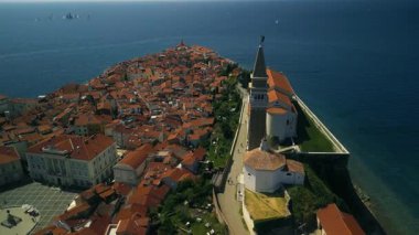 Çan kulesine sahip beyaz kilise, Piran şehri iç kısımlara doğru genişledikçe kayalık noktayı taçlandırıyor. Adriyatik Denizi uzakta parlarken, turuncu çatılar kıyı şeridi boyunca uzanıyor