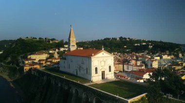 Yüksek çan kulesi olan beyaz kilise kümelenmiş eski evlerin tepesinde duruyor. Adriyatik Denizi gün ışığında parlarken Horizon kıyı şeridinde genişliyor