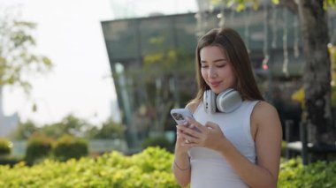 Boynunda kulaklık olan gülümseyen kadın portresi Green Park 'ta akıllı telefon tutuyor. Mutlu bayan internete akıllı telefonuyla bakıyor. Şehir merkezindeki yemyeşil çalılara karşı duruyor.