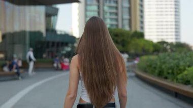 Uzun kahverengi saçlı zayıf genç bir kadın şehir merkezinin arka manzarası boyunca yürüyor. Kadın, yüksek binalara karşı sağlıklı doğal saç stili gösteriyor.