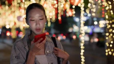 Güzel Asyalı kadın portresi şehir merkezindeki parlak bir gecede akıllı telefondan sesli mesaj kaydediyor. Gülümseyen Çinli kadın cep telefonuyla konuşuyor. Büyük akşam şehrinde eller serbest moda giriyor.
