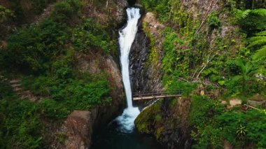 Endonezya 'nın Bali kentindeki yemyeşil alanda çağlayan bir şelalenin havadan görünüşü. Kırsal ahşap bir köprü akarsuyun üzerinden geçerek huzurlu bir atmosfer yaratıyor..