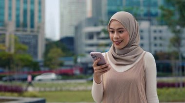 Müslüman kadın portresi, şehir manzarasına karşı yapılan akıllı telefondan zafer haberlerini okuyor. Geleneksel türbanlı Malezyalı kadın modern şehirde çevrimiçi piyangoda kazandı