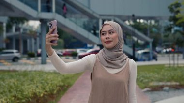 Genç Malezyalı tesettürlü kadın portresi şehir ortamında akıllı telefon standlarıyla selfie çekiyor. Güzel Müslüman kadın gülümseyerek günlük hayatı yakalar. Sosyal medya