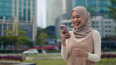 Başörtüsü takan genç iş kadınının portresi şehir merkezindeki akıllı telefon standlarında heyecan verici haberler okuyor. Malezyalı kadın şehir sokaklarında şaşkınlık ve mutluluğun karışımını ifade ediyor