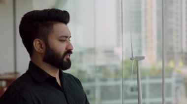 Düşünceli Hintli işadamının portresi ofis penceresinin yanındaki minyatür rüzgar türbinine bakıyor. Sakallı mühendis güç yapısına yeşil teknoloji entegrasyonu planlıyor