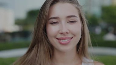 Şehir parkında rüzgarda dalgalanan saçlarıyla gülümseyen sakin genç kadın portresi. Güzel bayan yaz kenti ortamında dururken olumlu duygular ifade ediyor. Gençlik güzelliği