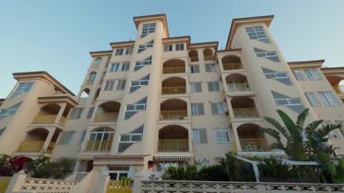Mavi gökyüzünün altında kemerli terasları ve düzensiz pencereleri olan modern bir otel binası. Şehir merkezinde palmiye ağaçları olan lüks bir pansiyon.