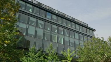 Ağaçlarla çevrili yansıtıcı cam cepheli modern ofis binası. Ticari büyüme, yenilik, sürdürülebilirlik ve kentsel kalkınmayı temsil eden temiz kurumsal mimari.