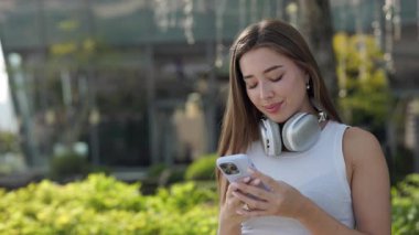 Kulaklıklı neşeli bir kadının portresi şehir parkında akıllı telefon kullanıyor. Gülümseyen kadın sosyal medyaya cep telefonuyla bakıyor ve şehir merkezindeki yeşil bitkilere karşı duruyor.