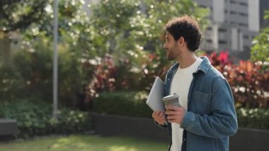 Dijital tabletli mutlu İspanyol adamın portresi yeşil şehir parkında kahve içiyor. Latin erkek öğrenci, şehir merkezinde bahar boyunca ağır çekimde yürürken mobil cihaz tutuyor.