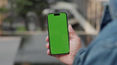 Modern şehirde reklam için yeşil renkli ekranlı akıllı telefon kullanan bir adamın yakın görüntüsü. Erkek, şehir merkezinde durduğu için cep telefonunun boş bir maketini gösteriyor.