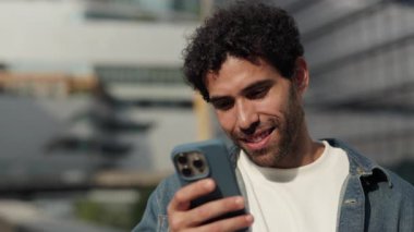 İspanyol adamın portresi akıllı telefondan mesaj okuyor ve Megapolis bloğunda gülümsüyor. Genç Latin Amerikalı erkek cevapları sosyal medyada güneşli bir şehirde cep telefonuyla yazdı.