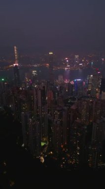 Yamaçlar yığılmış kulelere ve parlak sokaklara inerken liman ötesinde parlıyor. Uzak tepe bulanık ve sabit pencere desenleri Hong Kong dikey videosu boyunca ızgara oluşturur