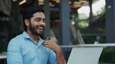 Kafedeki dizüstü bilgisayarla işaret diliyle konuşan gülümseyen Hintli girişimci portresi. Dost canlısı bir adam çevrimiçi şirket ofisinde konuşurken jestler kullanır.