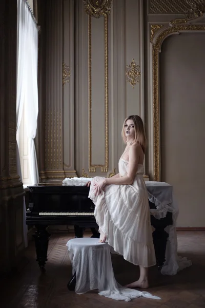 Bella ragazza in abito bianco vintage e un pianoforte in interni ricchi . — Foto Stock