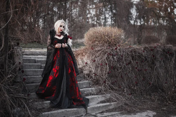 Mulher bonita com cabelo loiro longo em vestido vermelho à moda antiga andando através do jardim morto . — Fotografia de Stock