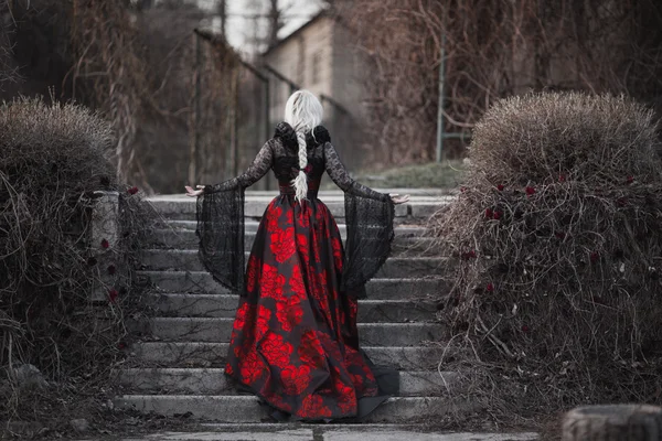 Mulher bonita com cabelo loiro longo em vestido vermelho à moda antiga andando através do jardim morto . — Fotografia de Stock