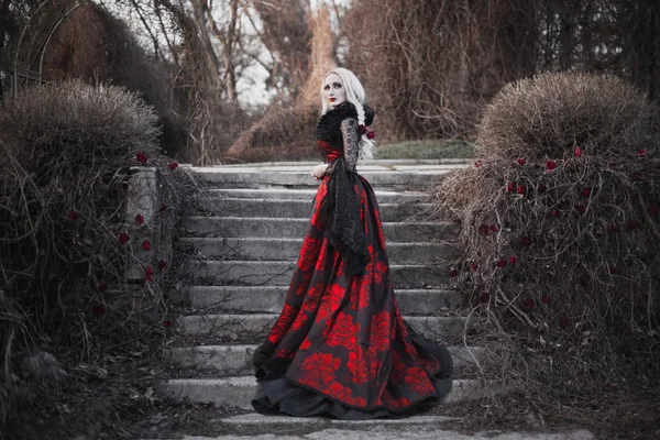 Hermosa mujer wBeautiful mujer con el pelo largo y rubio en vestido rojo pasado de moda caminando a través de muertos garden.ith pelo largo y rubio en vestido rojo pasado de moda — Foto de Stock