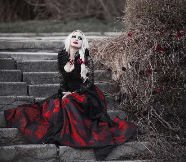 Belle femme wBelle femme avec de longs cheveux blonds dans une robe rouge à l'ancienne marchant à travers les cheveux blonds morts garden.ith longue robe rouge à l'ancienne — Photo