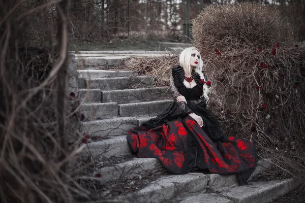 Hermosa mujer wBeautiful mujer con el pelo largo y rubio en vestido rojo pasado de moda caminando a través de muertos garden.ith pelo largo y rubio en vestido rojo pasado de moda — Foto de Stock