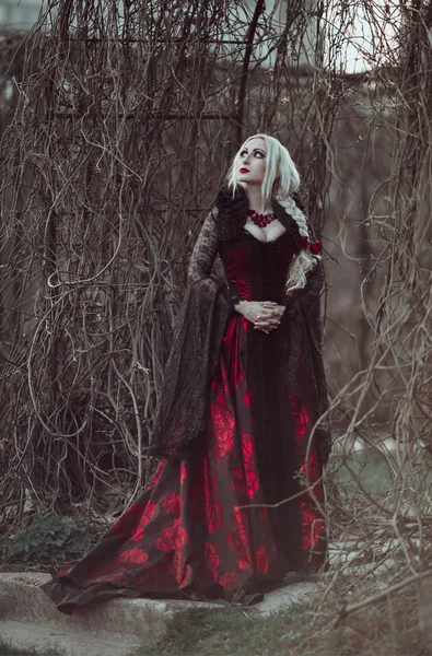 Belle femme wBelle femme avec de longs cheveux blonds dans une robe rouge à l'ancienne marchant à travers les cheveux blonds morts garden.ith longue robe rouge à l'ancienne — Photo