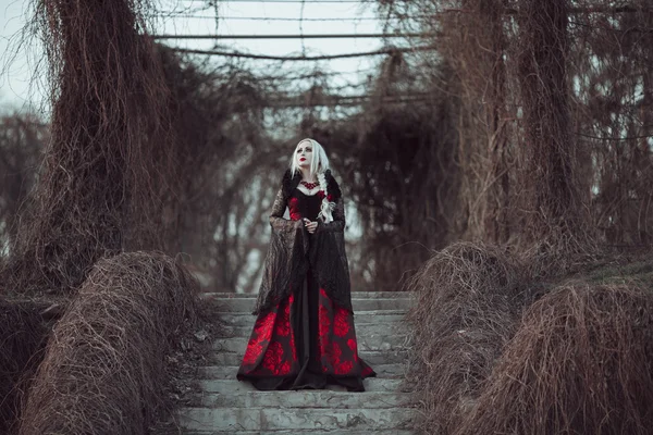 Женщина с длинными светлыми волосами в старомодном красном платье ходит по мертвому саду. — стоковое фото
