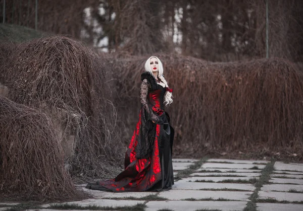 Bella donna wBeautiful donna con lunghi capelli biondi in vecchio vestito rosso stile a piedi attraverso morti garden.ith lunghi capelli biondi in vecchio vestito rosso stile — Foto Stock