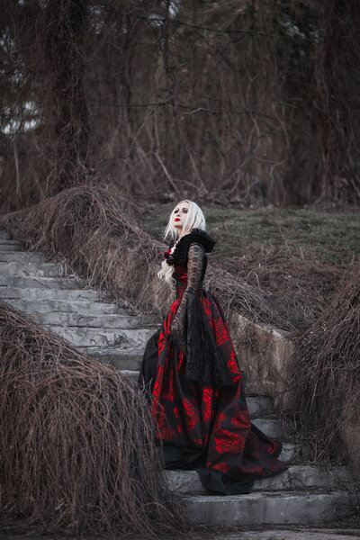 Beautiful woman with long blonde hair in old fashioned red dress walking through dead garden.
