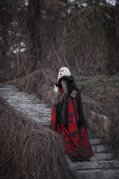 Женщина с длинными светлыми волосами в старомодном красном платье ходит по мертвому саду. — стоковое фото