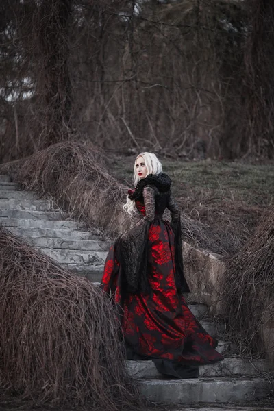 Schöne Frau mit langen blonden Haaren in altmodischem roten Kleid, die durch toten Garten geht. — Stockfoto