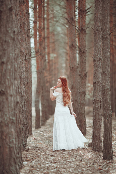 Ginger girl in white dress walking in pine forest.