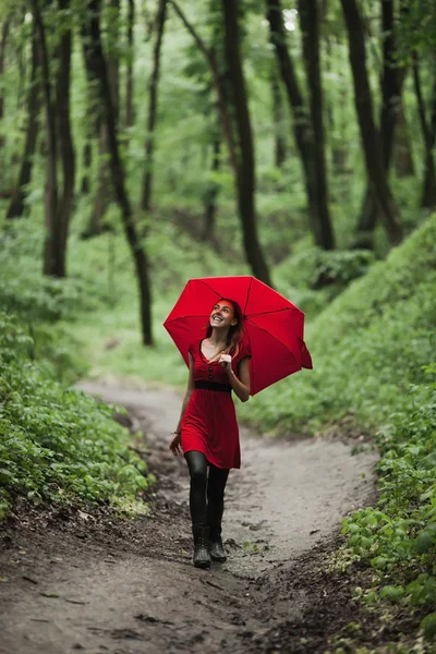 Happy girl in red dress walking in fresh green forest with umbrella. Rain concept. — Stock Photo, Image