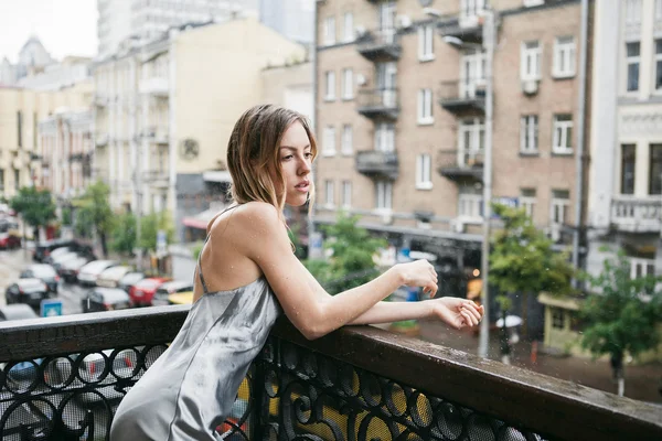 Mooi jong meisje in backless satijnen jurk enjoyng de regen op het balkon. — Stockfoto