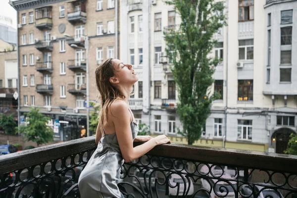 Mooi jong meisje in backless satijnen jurk enjoyng de regen op het balkon. — Stockfoto