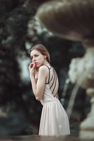 Hermosa joven en vestido rosa de moda de pie cerca de la fuente de piedra vintage . — Foto de Stock